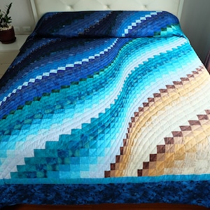May include: A quilted bedspread with a wave-like pattern in shades of blue, teal, and tan. The design features a geometric, stepped pattern, creating a textured appearance. The bedspread is on a bed with a white headboard.