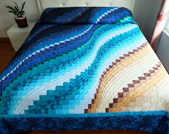 May include: A quilted bedspread with a wave-like pattern in shades of blue, teal, and tan. The design features a geometric, stepped pattern, creating a textured appearance. The bedspread is on a bed with a white headboard.