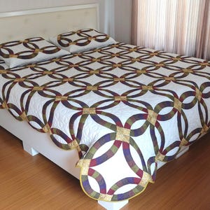 May include: A white bedspread and two pillowcases with a repeating pattern of interlocking circles in burgundy, olive green, and gold. The bed is on a white bed frame with a white headboard.