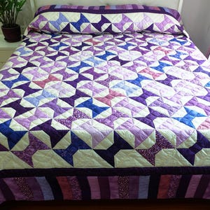 Op de afbeelding: Een handgemaakte quilt met een sterrenpatroon in tinten paars, lavendel en blauw. De quilt ligt op een bed, met een bijpassende kussensloop. De rand van de quilt is donkerder paars met gestreepte accenten.