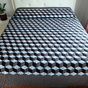 May include: A bedspread with a geometric cube pattern in black, gray, and white covers a bed. The quilt has a dark gray border and is neatly arranged on the bed. A small potted plant sits on a white nightstand.