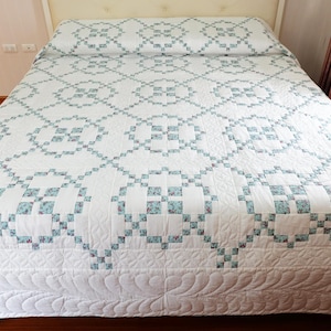 May include: A white quilt with a repeating pattern of blue and white squares, covering a bed. The quilt has a floral design within the squares and a decorative border. The bed is made up and ready for use.