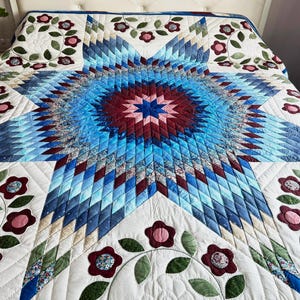 Könnte beinhalten: Handgefertigte Steppdecke mit einem zentralen Sternmuster in Blau-, Rot- und Weißtönen. Florale Applikationen in Rot und Grün zieren den weißen Hintergrund. Die Steppdecke wird auf einem Bett präsentiert und hebt ihr detailliertes Design hervor.