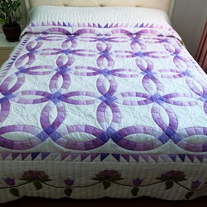 Op de afbeelding: Een handgemaakte quilt met een witte achtergrond en een herhalend patroon van in elkaar grijpende paarse ringen. De quilt heeft een bloemenrand en wordt weergegeven op een bed met een wit hoofdbord.
