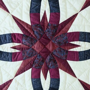 May include: Close-up of a quilted textile featuring a starburst design. The quilt showcases a cream-colored background with a pattern of intersecting arcs and star points in shades of burgundy, navy, and patterned fabrics. The stitching is visible.