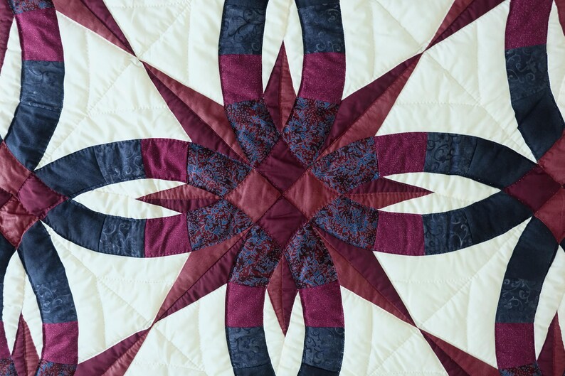 May include: Close-up of a quilted textile featuring a starburst design. The quilt showcases a cream-colored background with a pattern of intersecting arcs and star points in shades of burgundy, navy, and patterned fabrics. The stitching is visible.