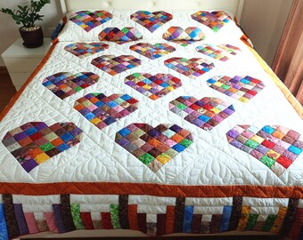 May include: A white quilt featuring a repeating pattern of multi-colored patchwork hearts. The quilt has a white background with intricate quilted stitching and a border composed of orange fabric and a vertical multi-colored block design.