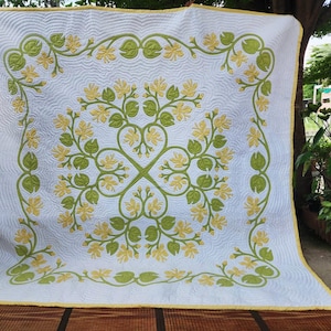 May include: A white quilt featuring a central symmetrical pattern of green vines and yellow flowers. The fabric has a wavy quilted texture throughout, and the entire piece is finished with a solid yellow border along all four edges.