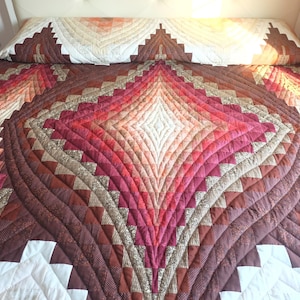May include: A quilted bedspread with a geometric design in shades of brown, red, and cream. The pattern features a central diamond shape with radiating lines and a repeating zigzag border. The bedspread is on a white bed.