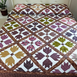 Puede incluir: Una cama cubierta con un edredón hecho a mano con un patrón repetido de huellas de oso estilizadas en tonos de marrón, morado, verde y dorado. El edredón está sobre una cama blanca con una almohada a juego y una falda de cama marrón. Una pequeña planta está en una mesita de noche blanca.
