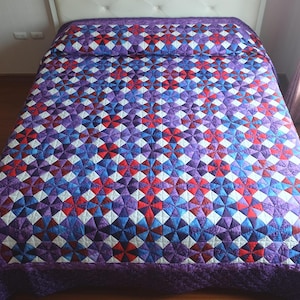 May include: A quilted bedspread with a geometric pattern in shades of purple, red, blue, and white. The bedspread is on a bed with a white headboard. The quilt has a patterned border.