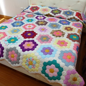 Pode incluir: Um edredom floral colorido com fundo branco. Apresenta um padrão repetido de flores hexagonais em várias cores, incluindo rosa, azul, amarelo e roxo. O edredom está em uma cama com cabeceira branca.