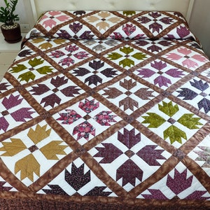 May include: A quilted bedspread with a repeating pattern of stylized bear paw prints in shades of brown, purple, green, and pink. The quilt is on a bed with a white headboard and a matching pillowcase. The bedspread has a brown border.