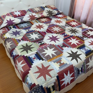 Op de afbeelding: Een patchwork sprei met een herhalend patroon van achtpuntige sterren en log cabin blokken. De sprei is opgebouwd uit diverse stoffen vierkanten in tinten rood, bruin, groen, paars en crème, die het oppervlak van een bed bedekken.
