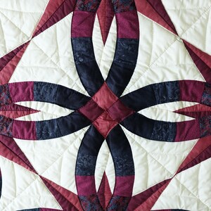 May include: Close-up of a quilted textile featuring a repeating pattern of interlocking rings and starburst shapes. The quilt showcases a color palette of cream, burgundy, and navy blue, with intricate stitching details. The design is symmetrical and geometric.