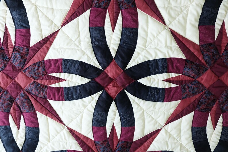 May include: Close-up of a quilted textile featuring a repeating pattern of interlocking rings and starburst shapes. The quilt showcases a color palette of cream, burgundy, and navy blue, with intricate stitching details. The design is symmetrical and geometric.