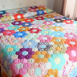 Pode incluir: Uma colcha colorida feita à mão com um padrão floral. A colcha apresenta um design repetitivo de formas de flores hexagonais em várias cores, incluindo rosa, azul, laranja e roxo. A colcha está em uma cama com uma cabeceira branca.