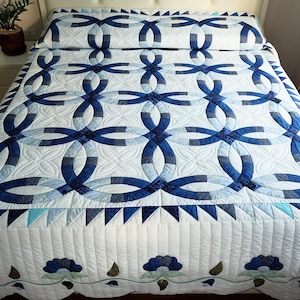 May include: A handmade quilt with a double wedding ring pattern in shades of blue and white. The quilt features a repeating design of interlocking rings, with a scalloped edge and floral accents. The quilt is on a bed with a matching pillow.