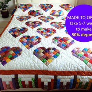 以下が含まれることがあります： 手作りのキルトで、ハート模様が繰り返されています。 各ハートは、カラフルな布の正方形で構成されています。 キルトは白い背景、オレンジ色のボーダー、模様のある下端があります。 テキスト「MADE TO ORDER」が表示されます。