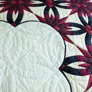 May include: A close-up of a quilt with a cream-colored background and a pattern of interlocking rings and starbursts in shades of burgundy and navy blue. The quilt has a white, quilted backing.