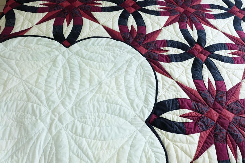 May include: A close-up of a quilt with a cream-colored background and a pattern of interlocking rings and starbursts in shades of burgundy and navy blue. The quilt has a white, quilted backing.