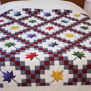 Puede incluir: Un edredón hecho a mano con un patrón repetitivo de cuadrados a cuadros rojos y blancos con aplicaciones de hojas de colores. Las hojas son de color rojo, amarillo, verde, azul y morado. El edredón está sobre una cama con una sábana blanca.