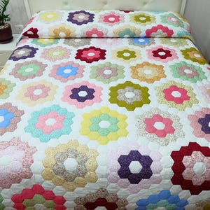 Op de afbeelding: Een kleurrijke quilt met een bloemenpatroon. De quilt heeft een witte achtergrond met talrijke zeshoekige bloemvormen in verschillende kleuren, waaronder roze, blauw, geel en groen. De quilt ligt op een bed met een wit hoofdbord.