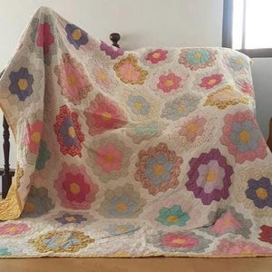 Puede incluir: Un edredón hecho a mano con un estampado floral en tonos de rosa, morado, azul y amarillo. El edredón está drapeado sobre un marco de cama y una escalera, con una guitarra en el fondo. El diseño del edredón presenta un patrón repetido de flores coloridas.