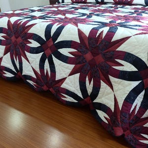 May include: A detailed shot of a bed covered with a quilt. The quilt features a repeating pattern of dark red and navy blue floral designs on a white background. The quilt is neatly arranged on the bed, showcasing the intricate stitching and design.