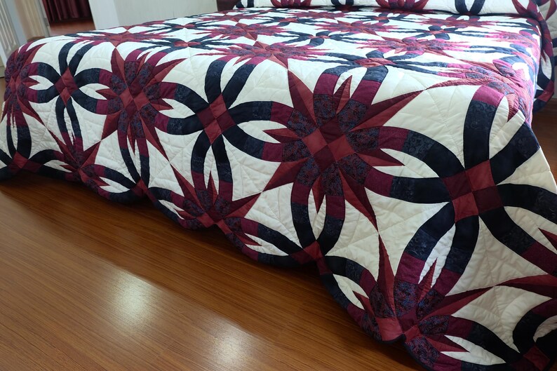 May include: A detailed shot of a bed covered with a quilt. The quilt features a repeating pattern of dark red and navy blue floral designs on a white background. The quilt is neatly arranged on the bed, showcasing the intricate stitching and design.
