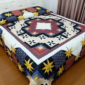 Può includere: Una trapunta colorata con un motivo geometrico e accenti di stelle e foglie. La trapunta presenta un motivo quadrato centrale in rosso, nero e beige, circondato da un bordo bianco. Stelle e foglie gialle sono sparse sulla trapunta.