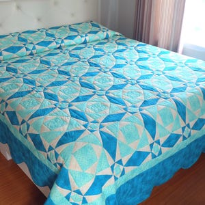 Könnte beinhalten: Ein Bett mit einer türkisfarbenen und weißen Steppdecke. Die Steppdecke hat ein geometrisches Muster aus Rauten und Sternen. Das Bett hat ein weißes Kopfteil und einen gewellten Rand an der Bordüre der Steppdecke.
