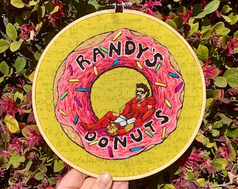 May include: Embroidered hoop art featuring a pink donut with sprinkles and a figure in a red suit sitting on top. The donut has the text "Randy's Donuts" written on it.
