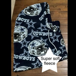 May include: Navy blue fleece balaclava featuring a repeating pattern of white Dallas Cowboys helmets, stars, and the team name. The text "Super soft fleece" is also visible. This balaclava is designed for warmth and comfort.