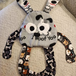 May include: A plush dog toy with a gray body, large white eyes, and black accents. The toy has floppy ears with dog faces and arms with a bone and paw print pattern. The text "I woof you" is printed on the front.