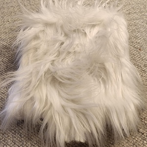 May include: A square, white faux fur rug with long, shaggy fibers. The rug has a soft, fluffy texture and is likely used for home decor.