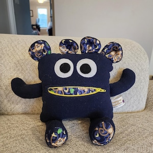 May include: A blue plush toy monster with a zipper mouth and monkey print ears and feet. The toy has large white eyes and a yellow zipper.