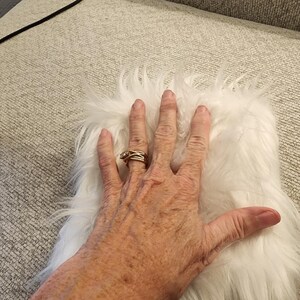 May include: A small, rectangular piece of white faux fur with long, soft fibers. The fur is fluffy and appears to be a solid color. The image shows the fur on a light gray surface.