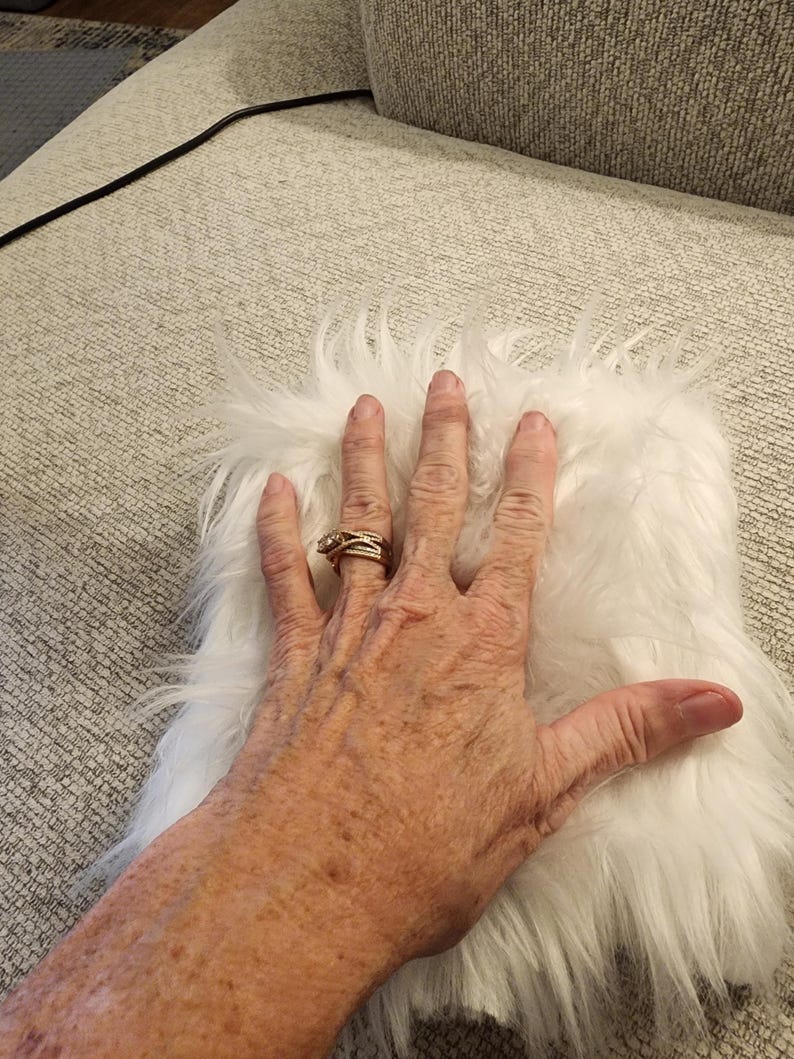 May include: A small, rectangular piece of white faux fur with long, soft fibers. The fur is fluffy and appears to be a solid color. The image shows the fur on a light gray surface.