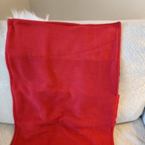 May include: A folded, solid red blanket with a soft texture. The blanket is resting on a light-colored couch with a white, fluffy trim visible at the top. The blanket's edges are neatly finished.