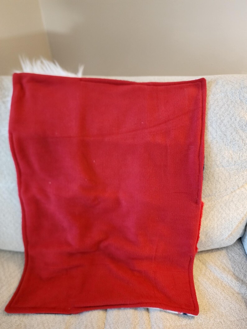 May include: A folded, solid red blanket with a soft texture. The blanket is resting on a light-colored couch with a white, fluffy trim visible at the top. The blanket's edges are neatly finished.