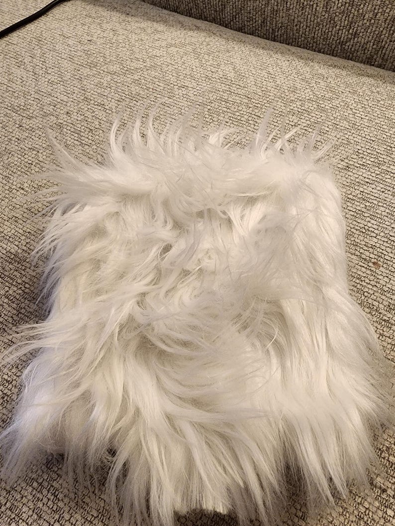 May include: A square, white faux fur rug with long, shaggy fibers. The rug has a soft, fluffy texture and is likely used for home decor.