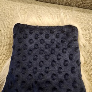 May include: A navy blue square pillow with a textured dot pattern. The pillow is surrounded by a fluffy white faux fur trim, creating a soft and inviting appearance. The pillow is likely for home decor.
