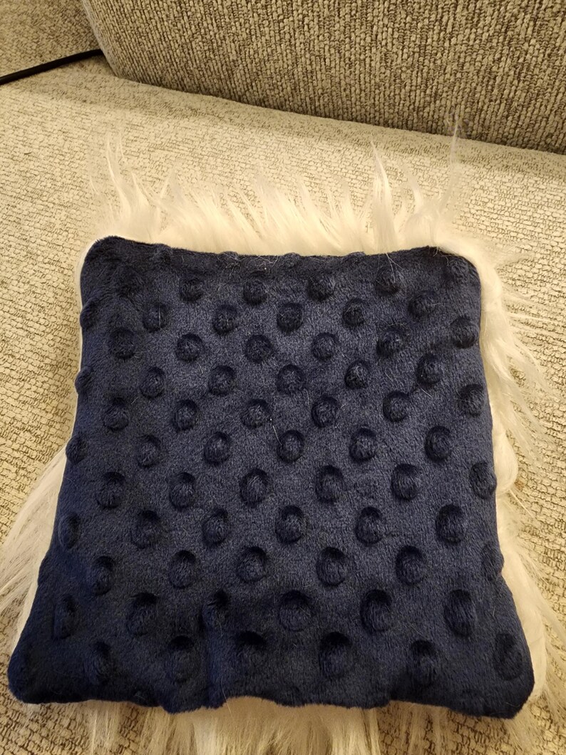 May include: A navy blue square pillow with a textured dot pattern. The pillow is surrounded by a fluffy white faux fur trim, creating a soft and inviting appearance. The pillow is likely for home decor.