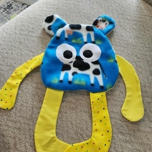 Op de afbeelding: Een blauw en geel pluche speelgoed met een koeienontwerp. Het speelgoed heeft een blauwe kop met grote witte ogen en een koeienillustratie. Het heeft gele armen en benen, waarbij de benen een stippenpatroon hebben. Kinderspeelgoed.