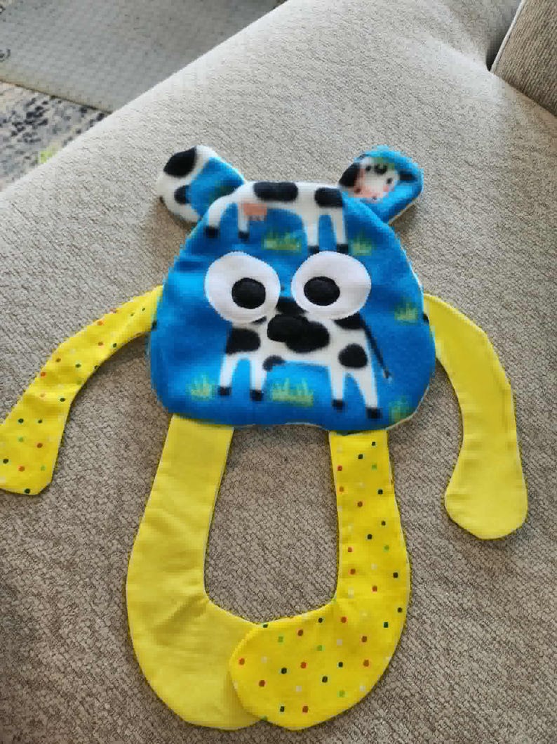 Op de afbeelding: Een blauw en geel pluche speelgoed met een koeienontwerp. Het speelgoed heeft een blauwe kop met grote witte ogen en een koeienillustratie. Het heeft gele armen en benen, waarbij de benen een stippenpatroon hebben. Kinderspeelgoed.