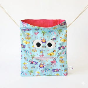 May include: A light blue drawstring bag featuring a repeating pattern of pink flamingos, palm trees, and beach umbrellas. The bag has two large white circular eyes with black pupils and a horizontal zippered mouth opening. A small tag reads Chomper Buddies.