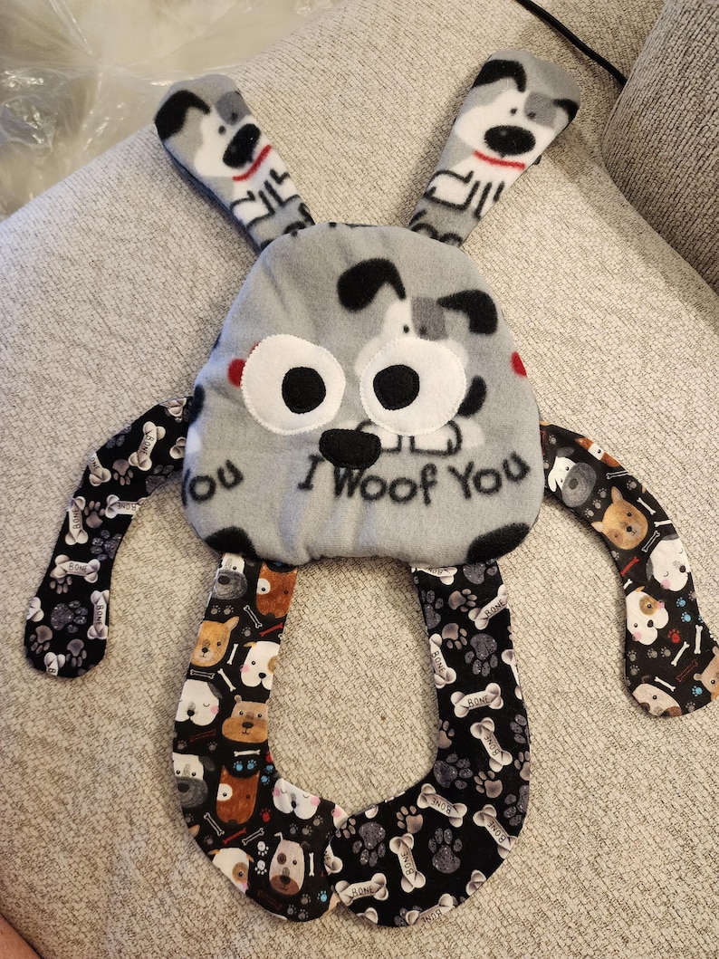 May include: A plush dog toy with a gray body, large white eyes, and black accents. The toy has floppy ears with dog faces and arms with a bone and paw print pattern. The text "I woof you" is printed on the front.