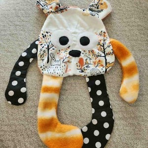 May include: A handmade plush toy in the shape of a bear with a whimsical design. The bear features a white face with large black eyes and a black nose. The arms are black with white polka dots, and the legs are orange and white striped. The ears have a floral pattern.