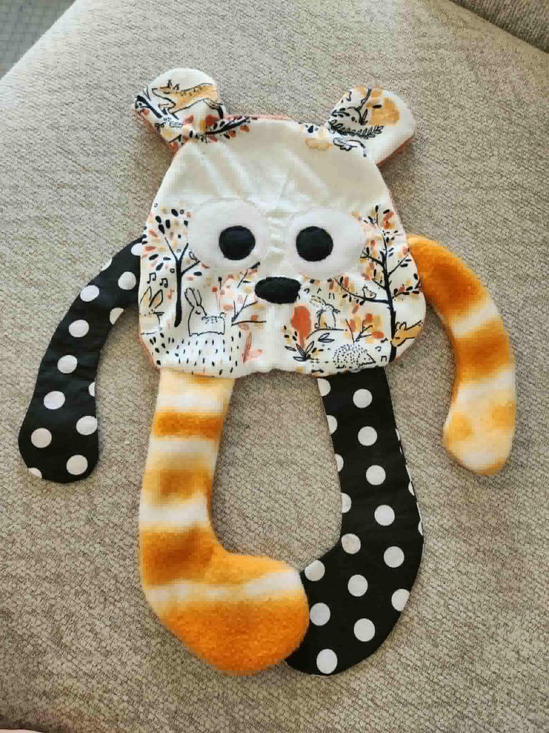 May include: A handmade plush toy in the shape of a bear with a whimsical design. The bear features a white face with large black eyes and a black nose. The arms are black with white polka dots, and the legs are orange and white striped. The ears have a floral pattern.